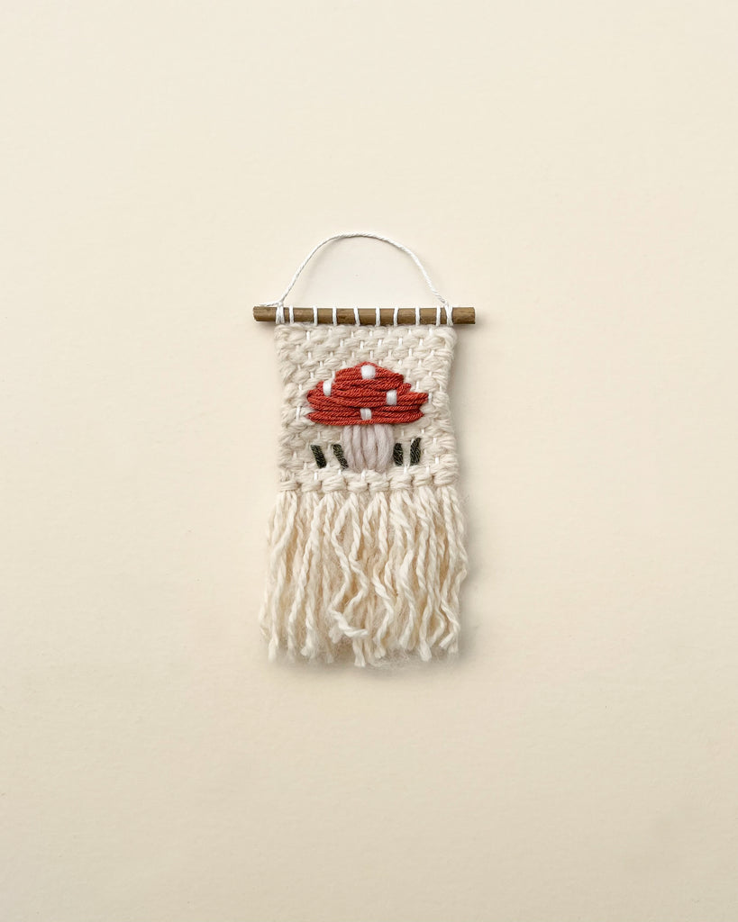 A handmade Mini Mushroom wall hanging featuring an embroidered representation of a red mushroom on a beige background, with white fringes hanging below, mounted on a horizontal stick.