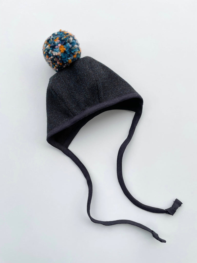 A Briar Baby Eon Pom Bonnet with ear flaps and a colorful blue and orange pompom on top, positioned on a plain white background.