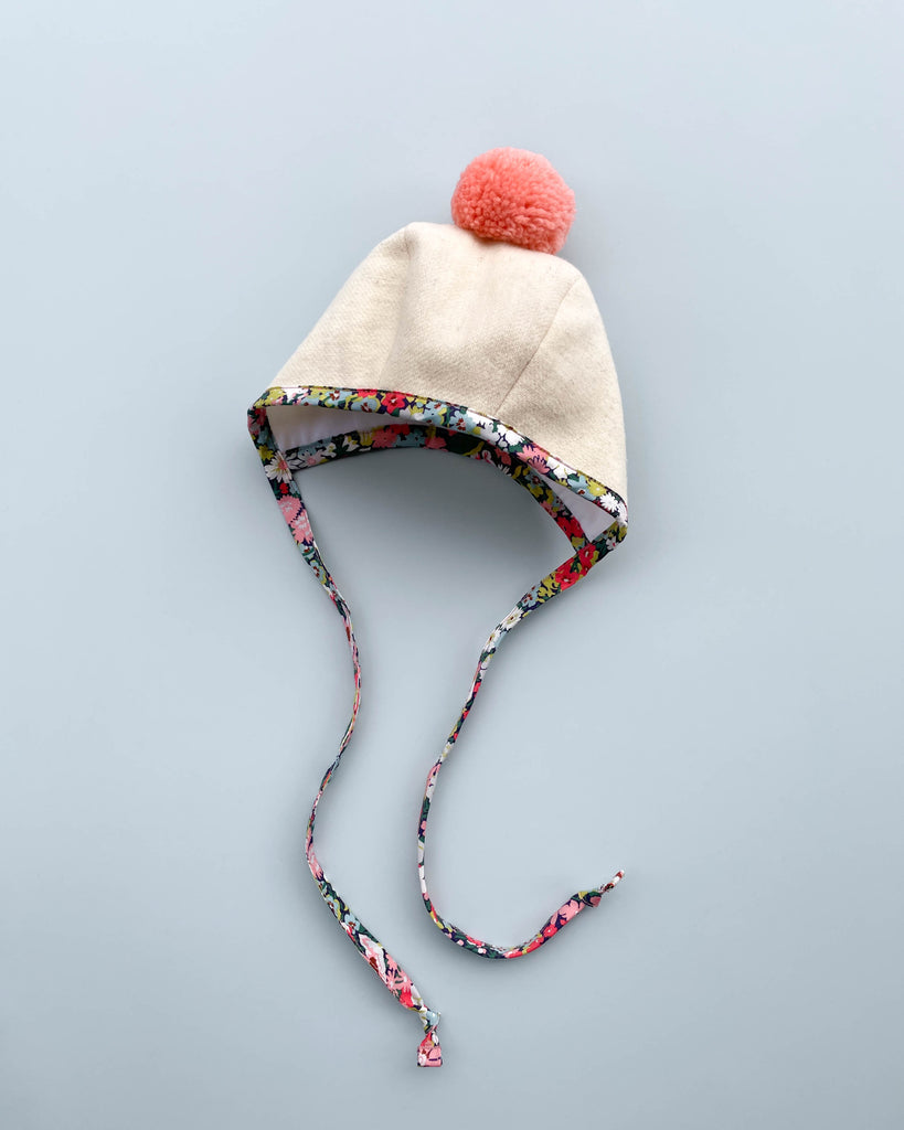 A Briar Baby Wild Poppy Pom Bonnet with a pink pompom on top and Liberty of London cotton floral trim, displayed against a plain gray background.