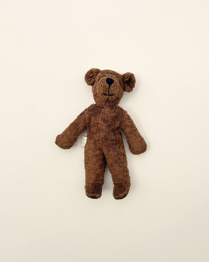 A small, fuzzy brown Senger Naturwelt Bear Stuffed Animal with black eyes and a subtle smile, expertly handmade by Senger Naturwelt and pinned vertically against a plain light beige wall.