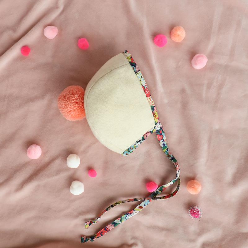 A Briar Baby Wild Poppy Pom Bonnet with a Liberty of London cotton ribbon lies on a pink fabric scattered with colorful pompoms. The image captures a playful and gentle aesthetic.