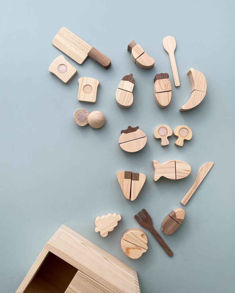 A collection of Velcro wooden vegetables and toy kitchen utensils, including forks, spoons, a wooden knife, and various fruits and vegetables, scattered on a light blue surface next to an open Hinoki