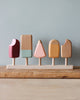 5 wooden popsicles on a stand in various popsicle shapes and in various pastel colors. Photographed against a light blue background.
