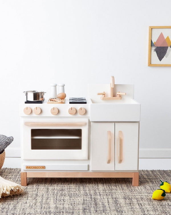 A Milton & Goose Wooden Play Kitchen - Made in USA, featuring a sustainable Baltic birch and white color scheme with a toy stove, oven, and sink, accessorized with pots, alongside a small wall hanging and plush