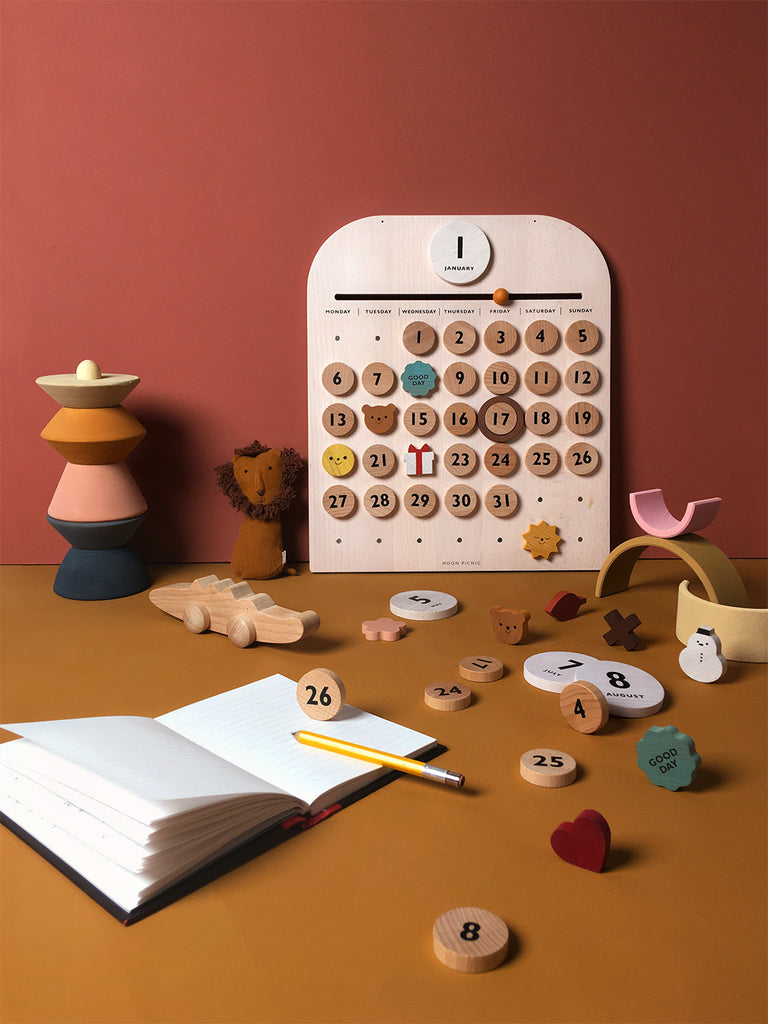 My Calendar featuring magnetic discs and showing March 1st, with scattered educational toys, a notebook, and a stack of colorful rings on a desk against a pink background.