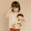 A young girl with a bob haircut is smiling and holding an Olli Ella Dinkum Doll. She is wearing a pastel pink top and rust-colored pants, standing against a beige background.