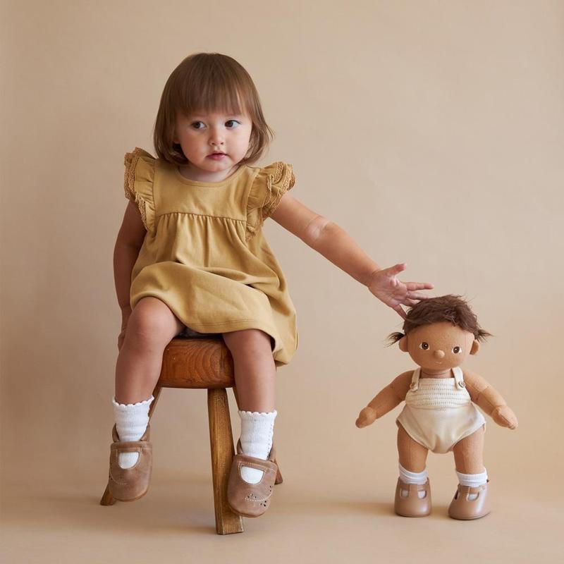 A young child in a Olli Ella Dinkum Dolls dress sits on a wooden stool, holding a posable doll beside her. She wears white socks and brown shoes, with a neutral background.