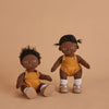 Two Olli Ella Dinkum Dolls with dark skin and black hair, dressed in yellow overalls, sitting against a beige background. One doll wears leg braces.