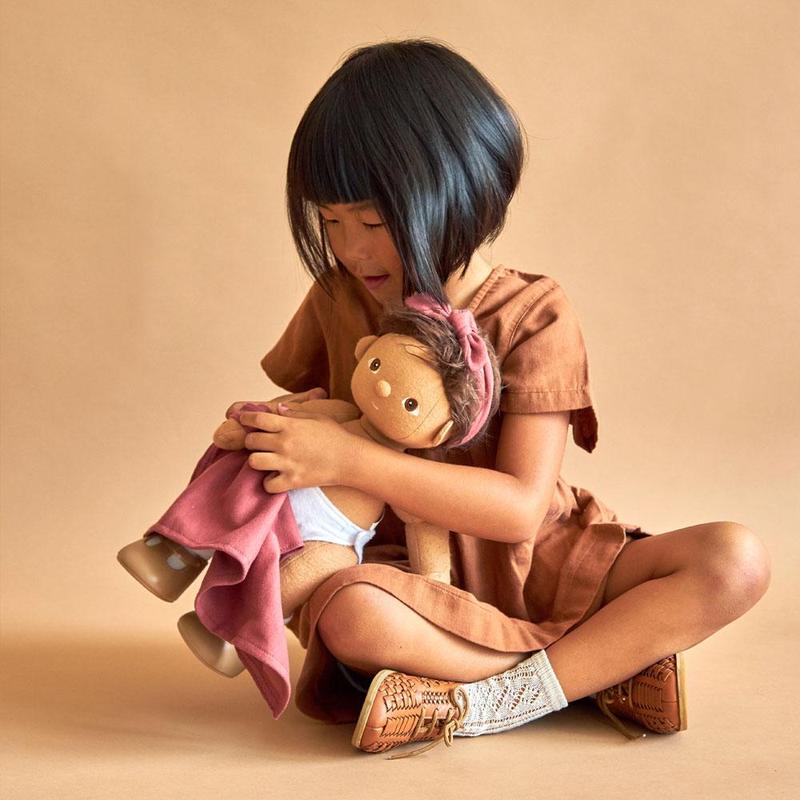 A young child with a bob haircut, wearing a brown dress, sits on the floor hugging an Olli Ella Dinkum Doll with affection.