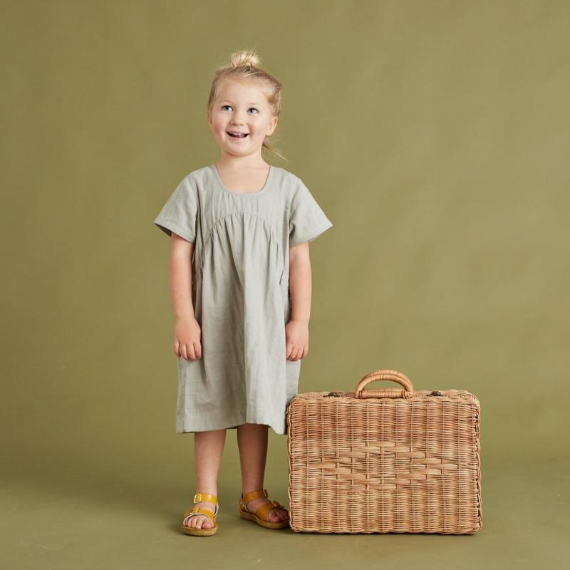 A young girl in a gray dress and yellow sandals smiles, standing next to a Rattan Toaty Trunk, on a green background.