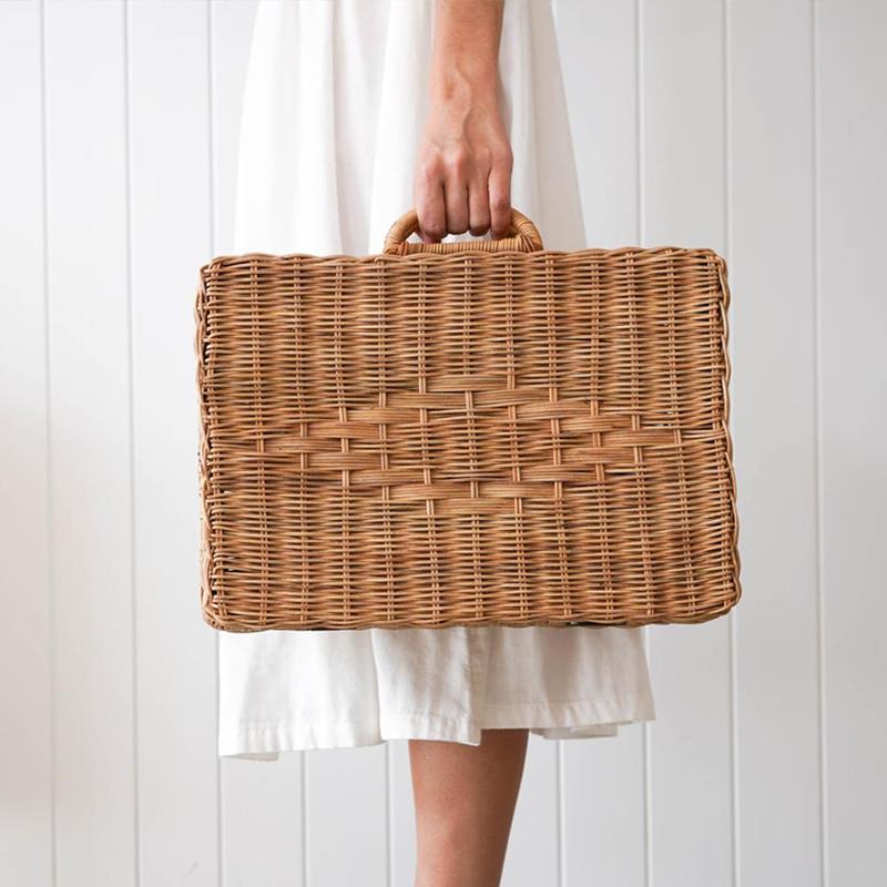 A person holding a natural Rattan Toaty Trunk in front of a white wooden panel background, with only their hand and lower half of a white dress visible.