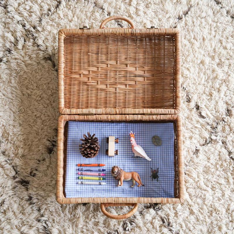 Sentence with product name: A Rattan Toaty Trunk open on a shaggy beige carpet, displaying items such as colored pencils, toy figurines, a pinecone, and a small chalkboard.