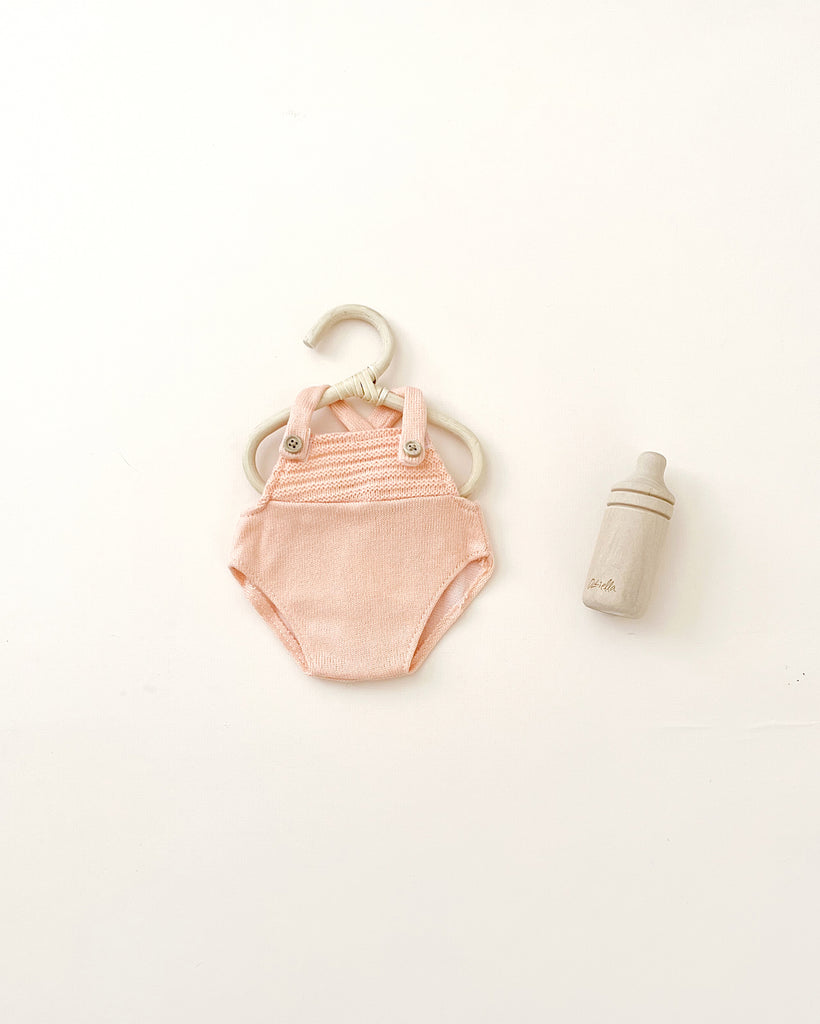 A soft pink cotton Olli Ella | Dinkum Doll Extra Clothing onesie accompanied by a small, light gray baby bottle, both items neatly placed on a plain, light background.