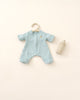 A baby's light blue cotton Olli Ella | Dinkum Doll Extra Clothing with a rainbow patch on a hanger next to a beige baby bottle, all displayed on a neutral background.