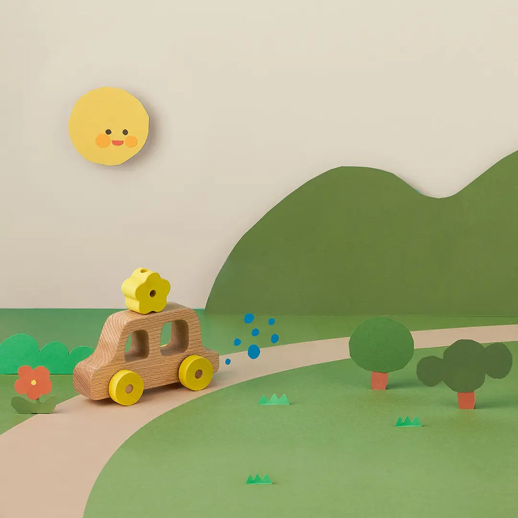 A playful scene with a My Little Car depicting a lion, driving on a green hill with stylized trees and flowers, under a smiling sun. Tiny blue dots represent movement behind the car.