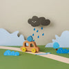 A crafted paper scene featuring a My Little Car with a blue sphere as the driver, under a frowning cloud raining blue drops, surrounded by paper bushes and waves on a green base.