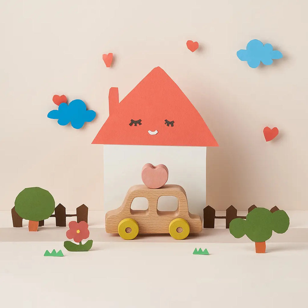 A playful scene made of beech wood and cut-out paper, depicting a house with a smiling face on its roof and My Little Car in front, surrounded by trees, clouds, and hearts.