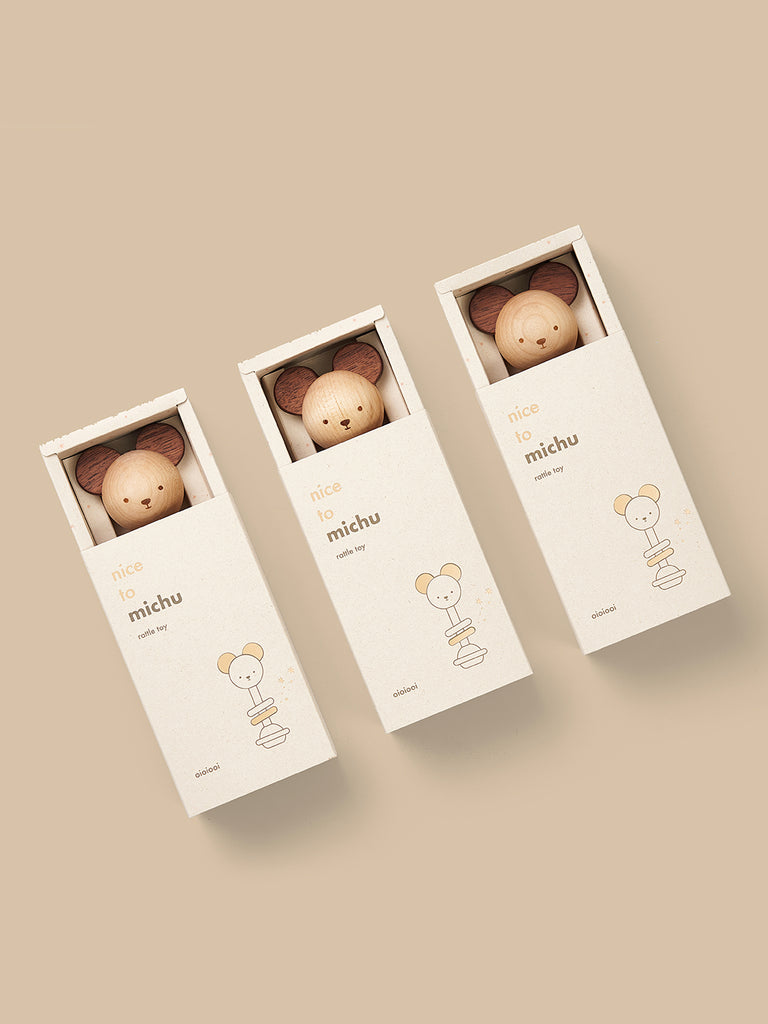 Three eco-friendly toy boxes labeled "Nice to Michu Baby Rattle" in a line, each containing a wooden smiling sphere designed to look like a character's head, displayed on a beige background.
