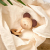 A Nice to Michu Baby Rattle shaped like a bear's face with large round ears, resting on a crinkled beige cloth with green leaves partially visible.