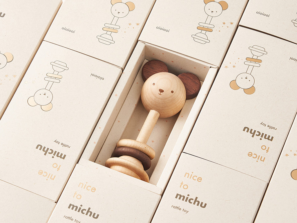 An eco-friendly Nice to Michu Baby Rattle displayed in an open cardboard box, with similar boxes featuring cute character illustrations lined up in the background.