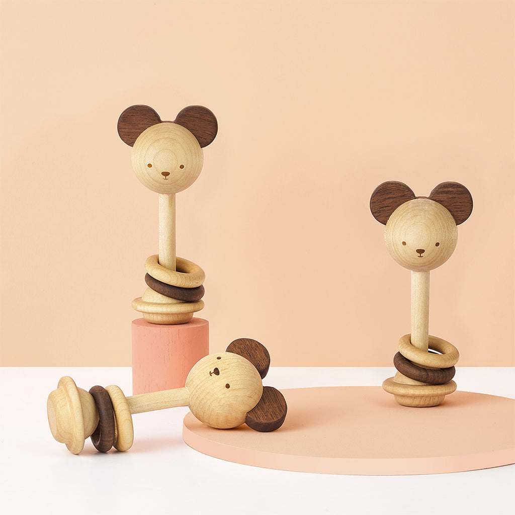 Two Nice to Michu eco-friendly wooden baby toys shaped like mice with circular heads and large ears, standing on soft pink and beige backgrounds. One toy features a stack of rings on a post, and the other a wheel.