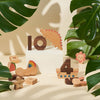 Numbers Play Block Set and toys, including a dinosaur and shapes, are arranged artistically among lush green monstera leaves on a neutral background.