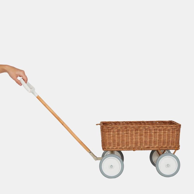 A hand holding the handle of an Olli Ella Rattan Wonder Wagon on wheels against a plain light background.