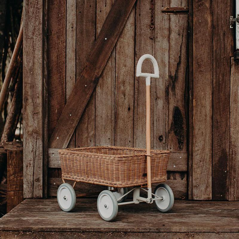 A vintage handmade Olli Ella Rattan Wonder Wagon on wheels with a handle, parked in front of an aged wooden barn door.