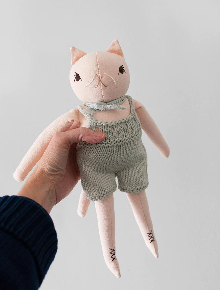 A hand holds a Polka Dot Club Cat in Hand Knit Overalls with hand embroidered features and dressed in baby alpaca overalls against a plain background.