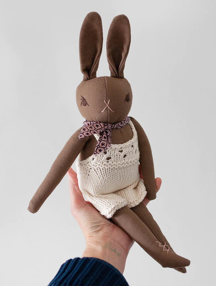 A hand holds up a Polka Dot Club Brown Rabbit in Hand Knit Overalls, featuring long brown ears, a stitched face, and dressed in baby alpaca overalls and a floral scarf.