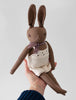A hand holds up a Polka Dot Club Brown Rabbit in Hand Knit Overalls, featuring long brown ears, a stitched face, and dressed in baby alpaca overalls and a floral scarf.