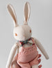 A hand holding a Polka Dot Club Cream Rabbit in Hand Knit Overalls with long ears and hand-embroidered features, wearing baby alpaca overalls and a floral scarf. The background is plain and light-colored.