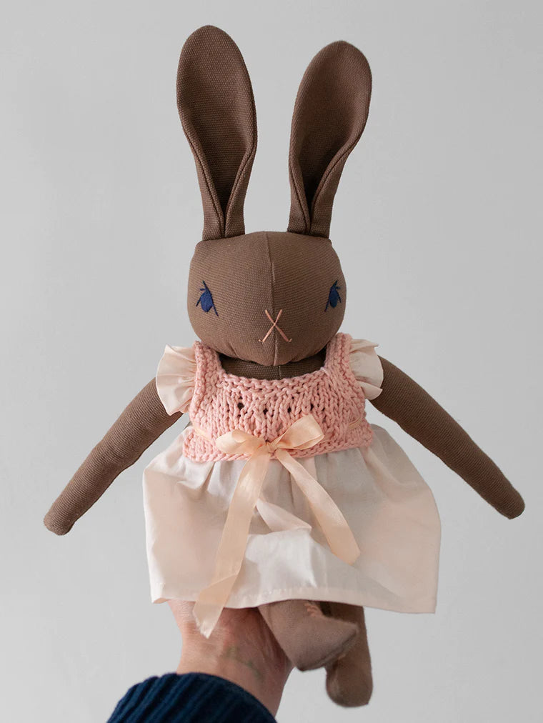 A hand holding a Polka Dot Club Large Brown Rabbit in Hand Knit Dress with brown ears and body. The bunny, crafted from cotton canvas, is dressed in a white dress with a pink knitted bodice adorned with a ribbon bow. The bunny has hand-embroidered blue eyes and nose details. The background is plain and neutral.