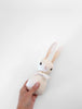 A hand holding a Polka Dot Club Rabbit Rattle - Peach with long ears, embroidered eyes, and an organic cream necktie around its neck against a white background.