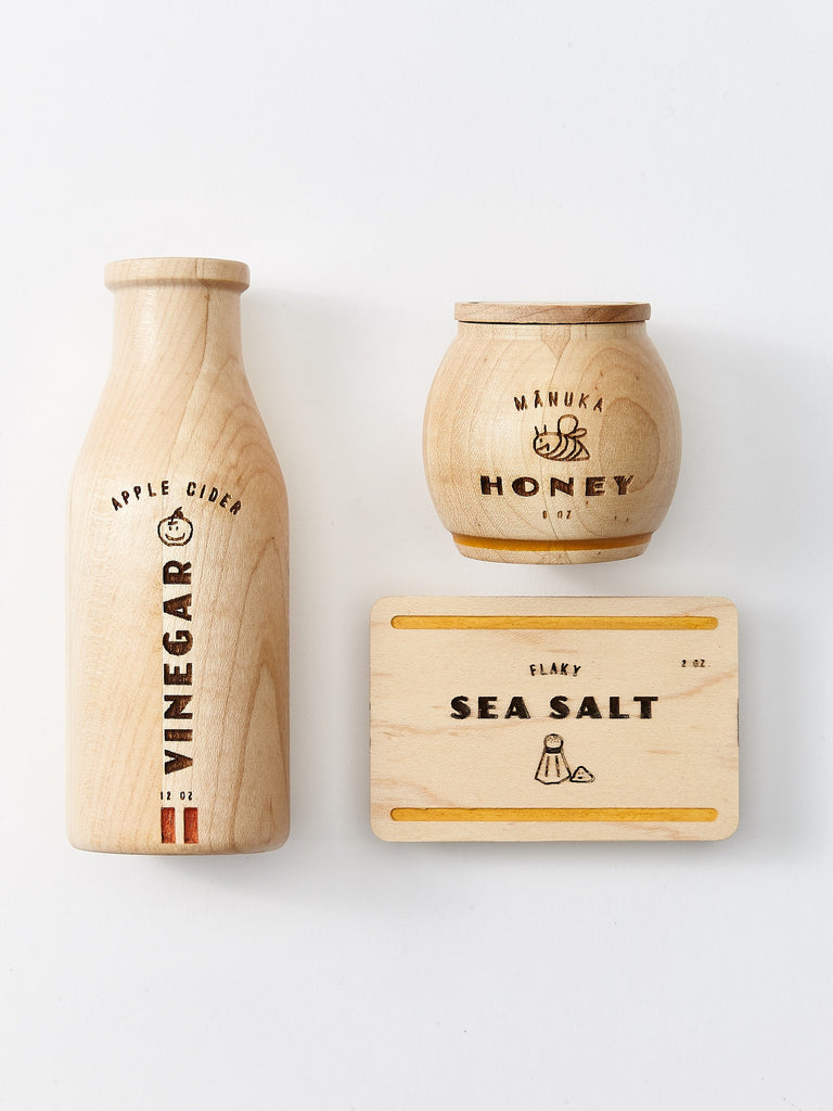 The Milton & Goose Pantry Play Food Set features a minimalist wooden design showcasing three items: a bottle labeled "Apple Cider Vinegar," a jar labeled "Manuka Honey," and a rectangular block of Flaky Sea Salt. Each item is adorned with simple, engraved black lettering and small illustrated imagery on a white background.