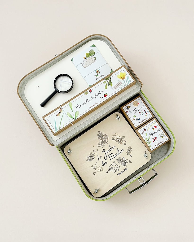 Sentence with product name: An open Moulin Roty The Botanist Suitcase-themed educational kit in a suitcase, featuring a magnifying glass, illustrated cards, and a wooden notebook with plant designs.
