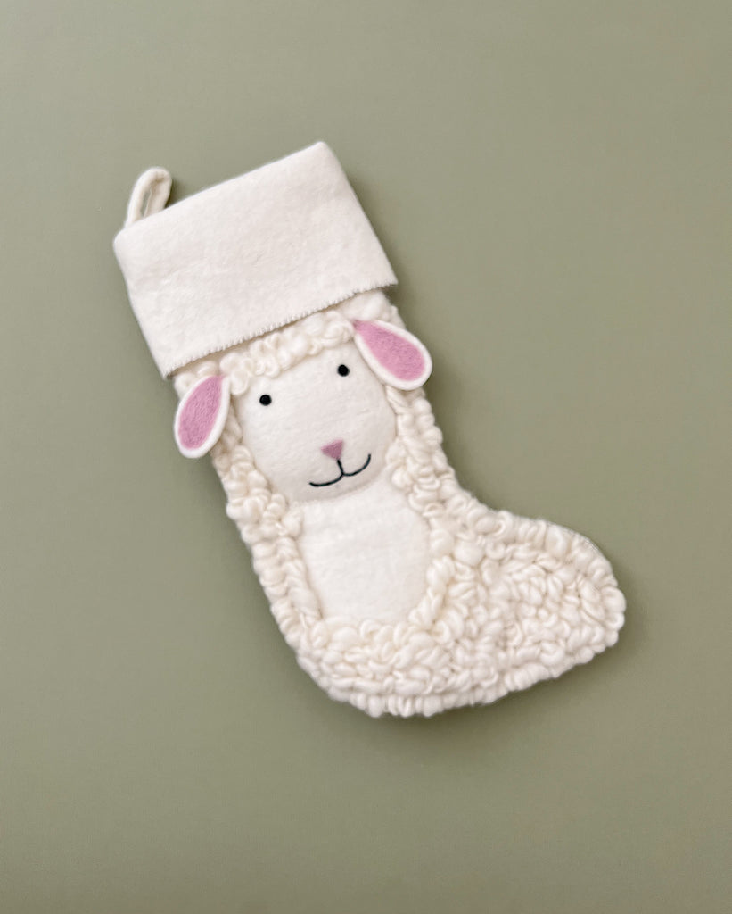 A soft white Handmade Sheep Christmas Stocking with a smiling face and pink ears, resembling a plush toy, hanging against a pale green background.