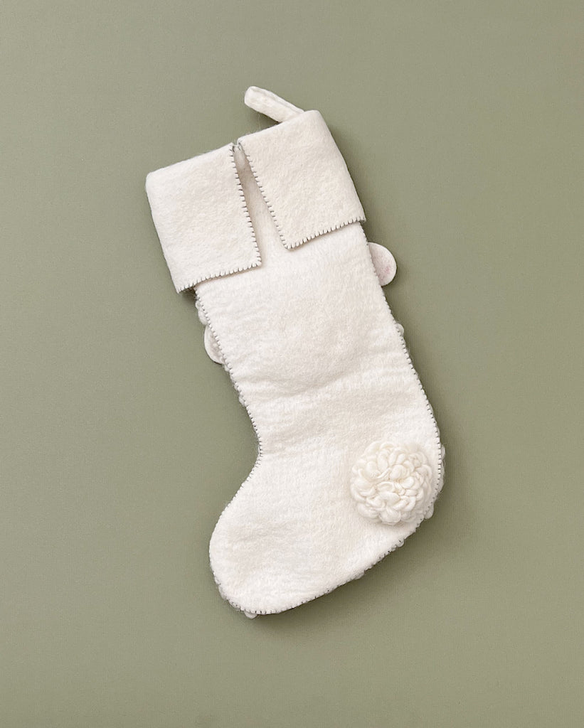 A Handmade Sheep Christmas Stocking made of soft organic felt is hanging against a plain, olive green background. The festive addition features a folded cuff and a decorative flower sewn near the toe area.
