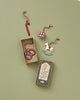 A set of vintage-style Maileg Peter's Christmas Ornament Set lies on a light green background. The decorations include a candy cane, a drum, and a rocking horse. A pretzel ornament is inside a small open gift box, with another box below featuring an illustration and text.