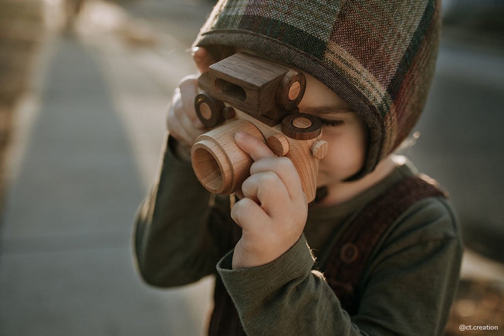 A child wearing a plaid hat plays with a Father’s Factory | Vintage Style Wooden Toy Camera, looking through its lens, with soft sunlight illuminating the scene.
