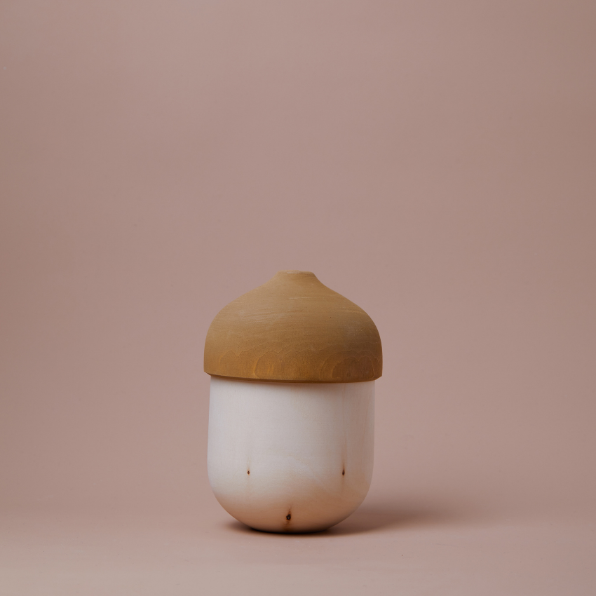 A set of Raduga Grez Nesting Acorns, crafted from solid wood with a smooth, round base and a slightly darker, rounded lid, set against a plain, light pink background.