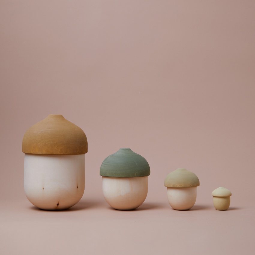 A set of four Raduga Grez Nesting Acorns with dome-shaped lids, decreasing in size, arranged in a row on a soft pink background. The largest has a tan lid, while the others are shades of green.