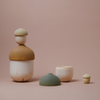 A collection of Raduga Grez Nesting Acorns, displayed against a soft pink background. The pieces feature natural wood tones and muted green accents.