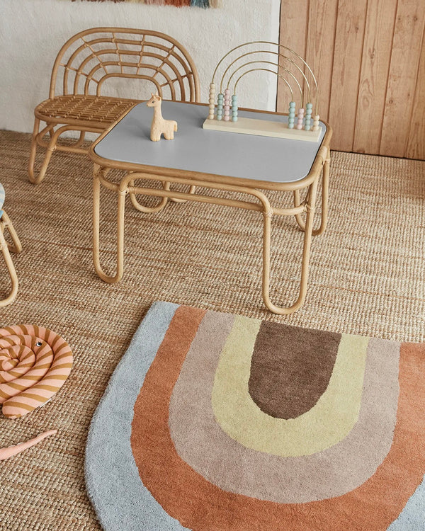 A cozy children's room decor featuring a rattan chair and table set on an Oyoy Rainbow Rug, with toys on the table and a patterned cushion nearby.