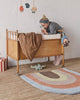A woman in a cozy sweater and headband peers into a vintage wooden crib with a baby inside, standing on an Oyoy Rainbow Rug in a warmly decorated room.