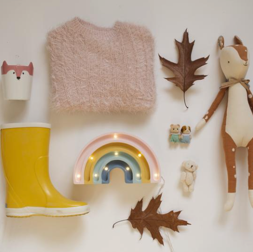Flat lay of children's items: a fluffy pink sweater, a yellow rubber boot, a Little Lights Mini Rainbow Lamp, two leaves, stuffed animal toys, and small decorative figures on a light