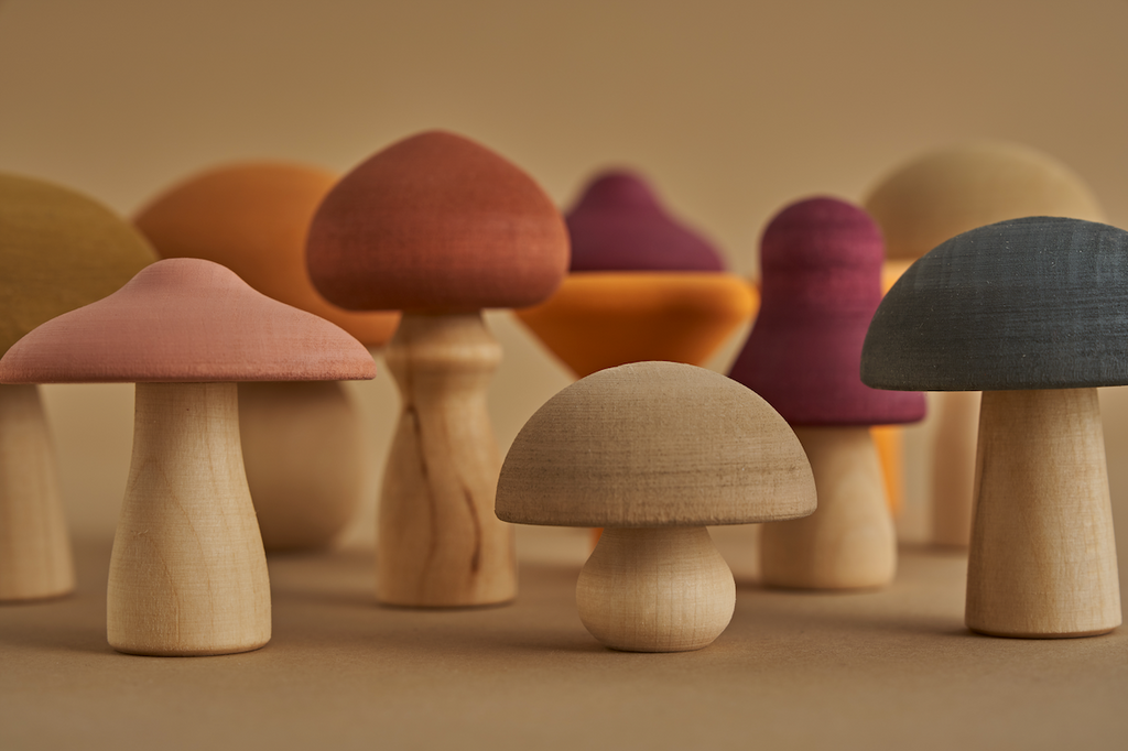 A collection of Raduga Grez Handmade Wooden Mushrooms in various shapes and earthy colors, crafted from wood with non-toxic paint, arranged against a beige background to highlight their natural tones and textures.