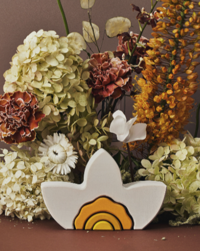 A colorful assortment of Raduga Grez Arch Stacker - Narcissus flowers, including hydrangeas, daisies, and other varieties, arranged around a small, bird-shaped ceramic vase on a brown background from a sustainability responsible supplier.
