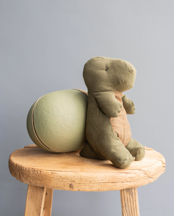 A plush toy hippopotamus sitting on a round wooden stool, viewed from behind, with a soft green finish and stitched details visible, resembling a natural looking Maileg | Medium Gantosaurus in egg - Green from the Gant