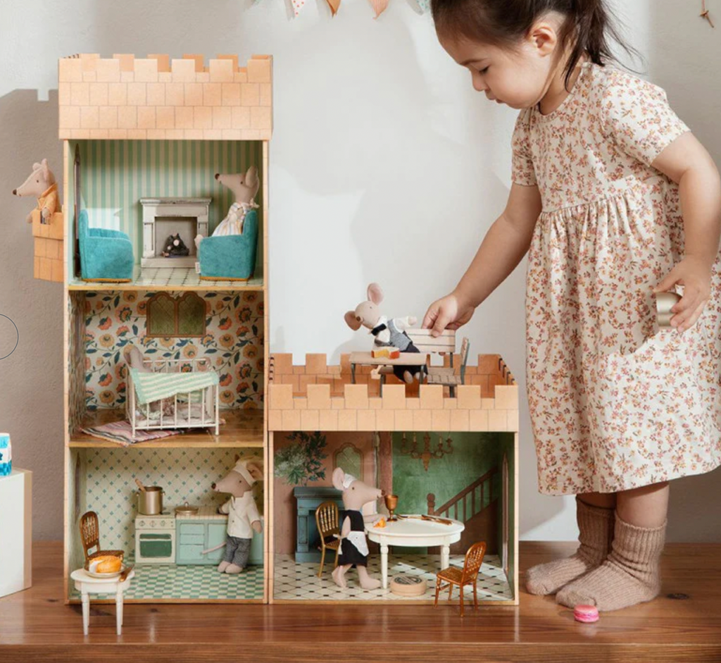 A young girl plays with a large, detailed Maileg Miniature Dining Table containing miniature furniture and animal figures, dressing in a floral dress and cozy socks.